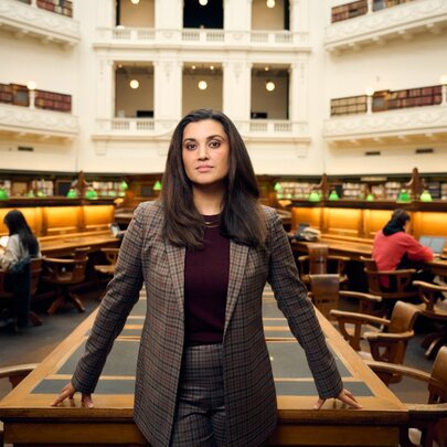 Host Jamila Rizvi, a woman wearing a tweed jacket standing in front of a desk in the State Library Victoria domed reading room.