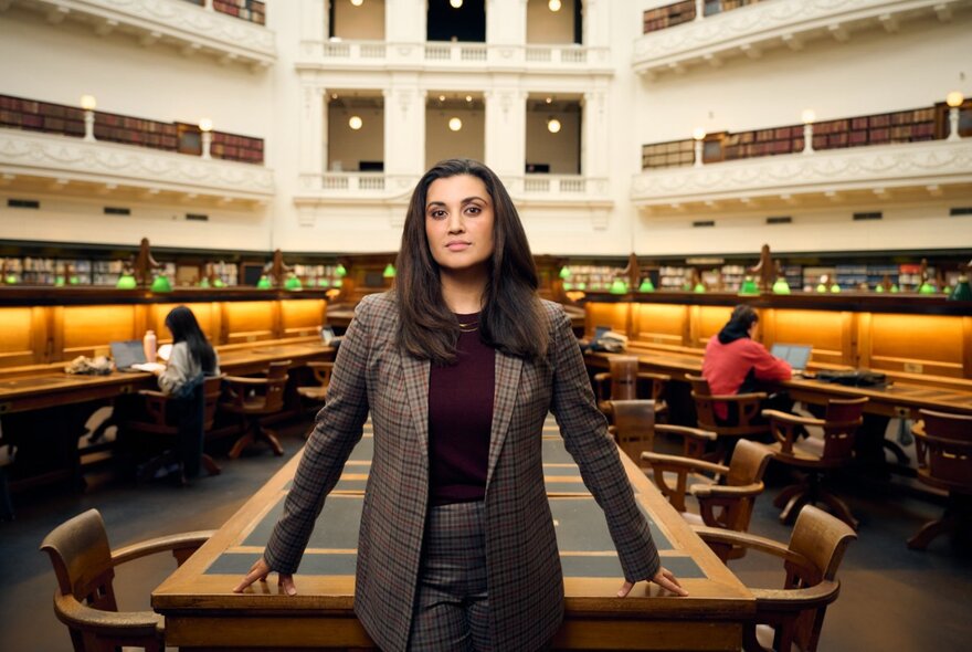 Host Jamila Rizvi, a woman wearing a tweed jacket standing in front of a desk in the State Library Victoria domed reading room.