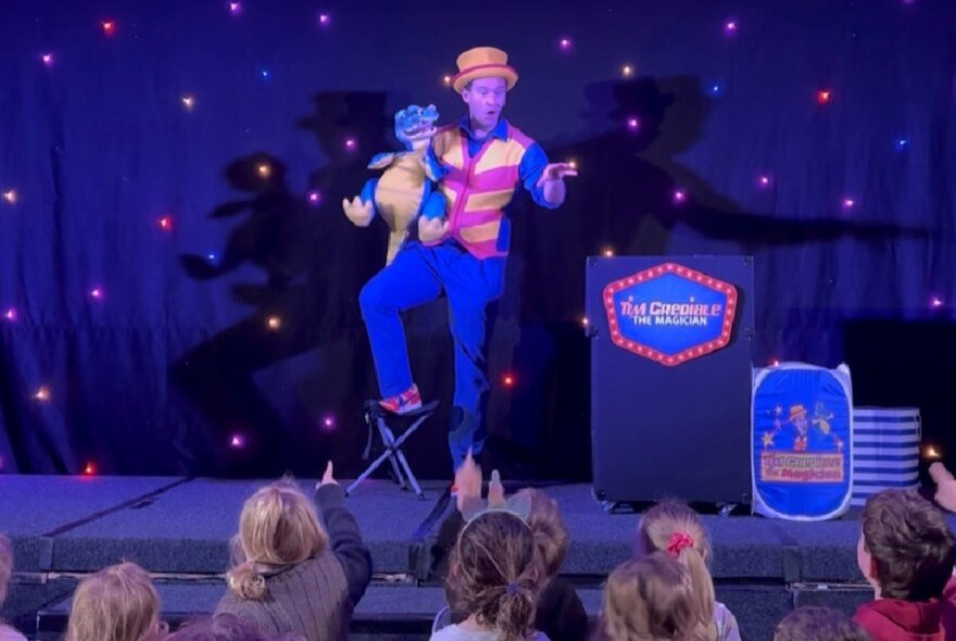 Magician Tim Credible on a dark star-lit stage with Douzie the Dragon puppet on his arm.