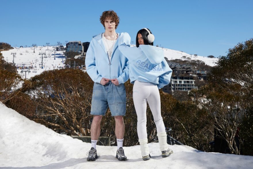 Two fashion models wearing jackets, shorts and leggings, standing in a snowy landscape.