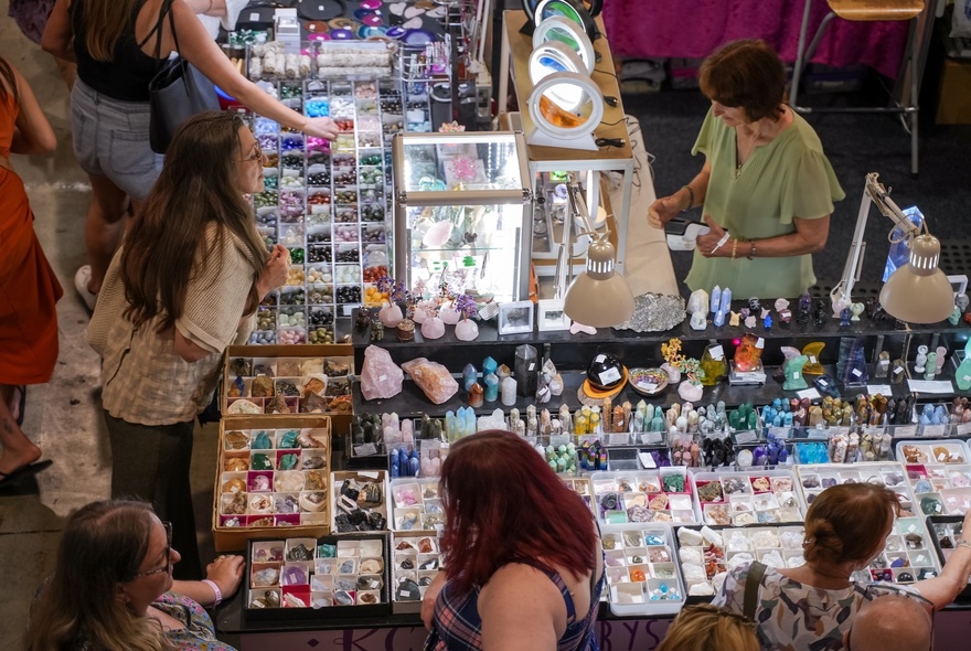 Overhead view of a bustling scene at a market stall, likely focused on crystals, gems, and handcrafted goods, with people browsing the stall.