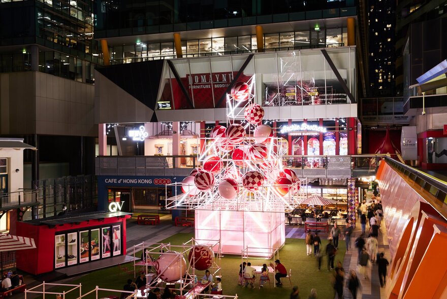 Christmas tree installation at QV square constructed from steel and oversized baubles at night, with people standing around, retail spaces in the background.