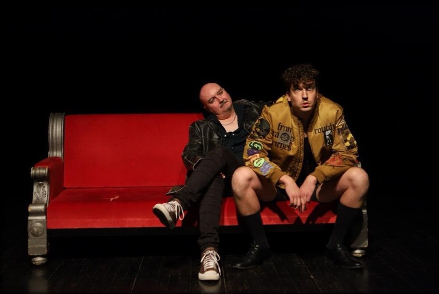 Two improv actors seated close to each other on a red bench seat on a stage.