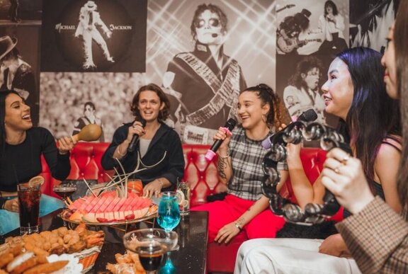 A group of friends singing in a private karaoke room, with the table in front of them filled with platters of food and drinks.