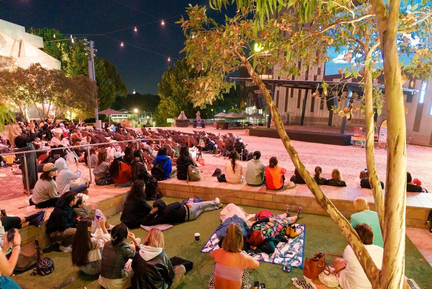 An audience sitting on the paved ground at Fed Square at night, watching a movie.