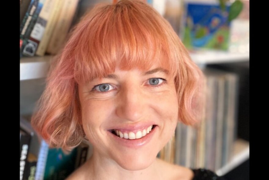 Smiling person with bob length pink hair, standing in front of a bookcase.