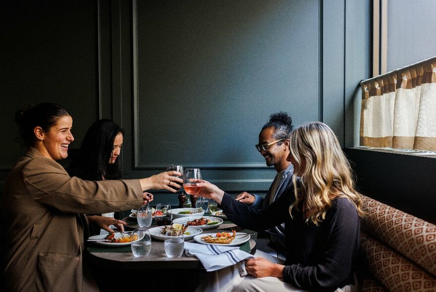 Diners seated at a table with banquet seating, toasting with raised glasses above dishes of food. 