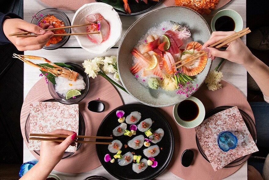 Hands with chopsticks selecting from bowls of sushi and sashimi.