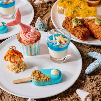 'Wonders of the sea' themed high tea treats including mermaid-tail cupcakes on a white plate on a display of sand and shells.