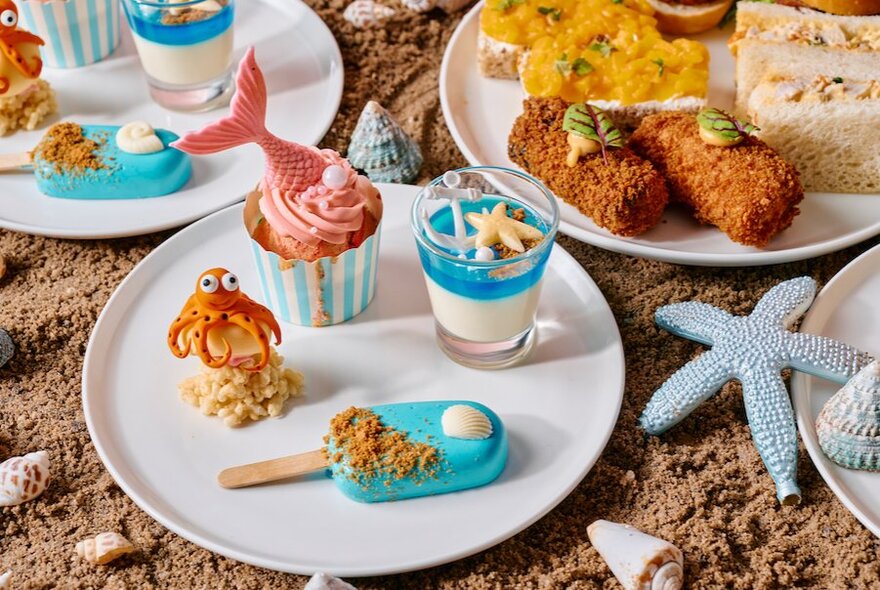 'Wonders of the sea' themed high tea treats including mermaid-tail cupcakes on a white plate on a display of sand and shells.