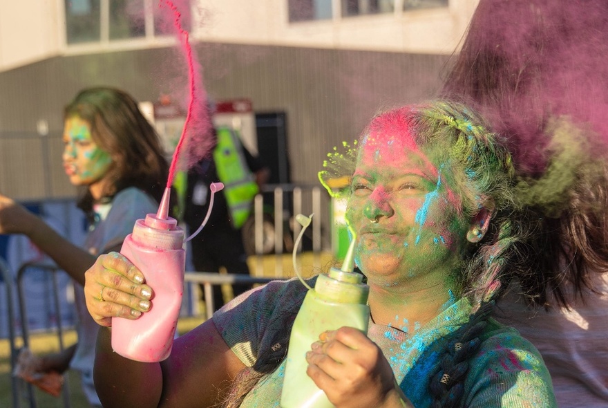 A person in a crowd celebrating and dancing, covered in coloured powder, and squirting coloured powder from squeezy bottles at Melbourne's outdoor Holi Festival.