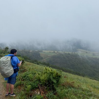 Hikers carrying large backpacks on a trail during the journey on the Camino de Santiago, with green hills and mist in the background.