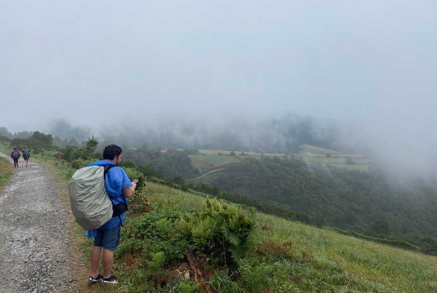 Hikers carrying large backpacks on a trail during the journey on the Camino de Santiago, with green hills and mist in the background.