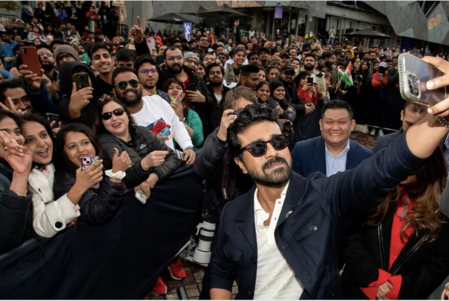 An Indian celebrity wearing dark sunglasses taking a selfie with a large crowd of cheering fans behind him; at Fed Square.