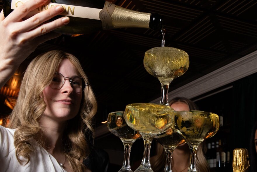 A bartender pours champagne into glasses arranged in a tower. 