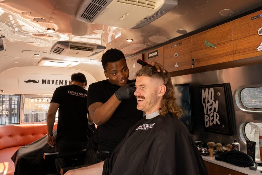 A long-haired man in a converted Airstream trailer, smiling as a barber shaves his hair into a mullet style.