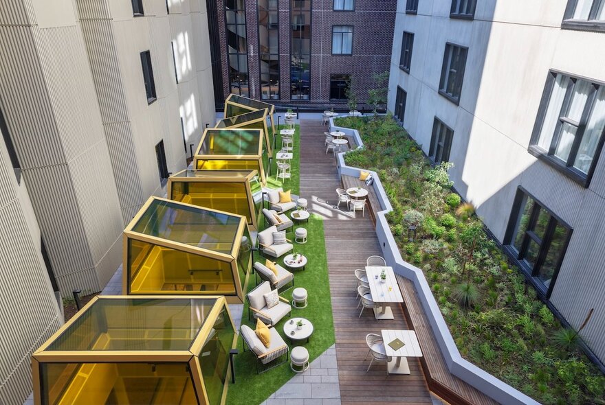 Looking down on the outdoor terrace and courtyard of a hotel from above, with greenery, occasional tables, chairs and large armchairs. 