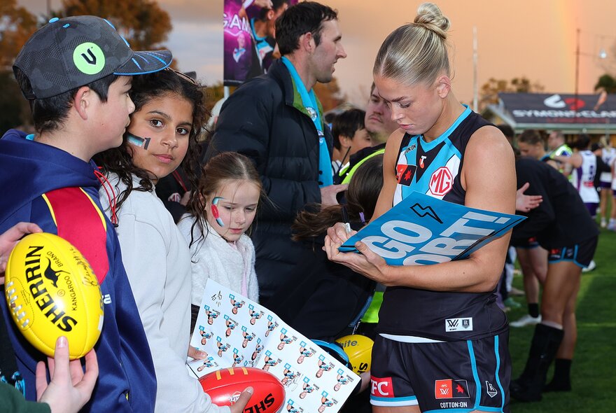 Young fans lining up in front of a Port Adelaide AFLW player with their footballs and caps, waiting for her signature.
