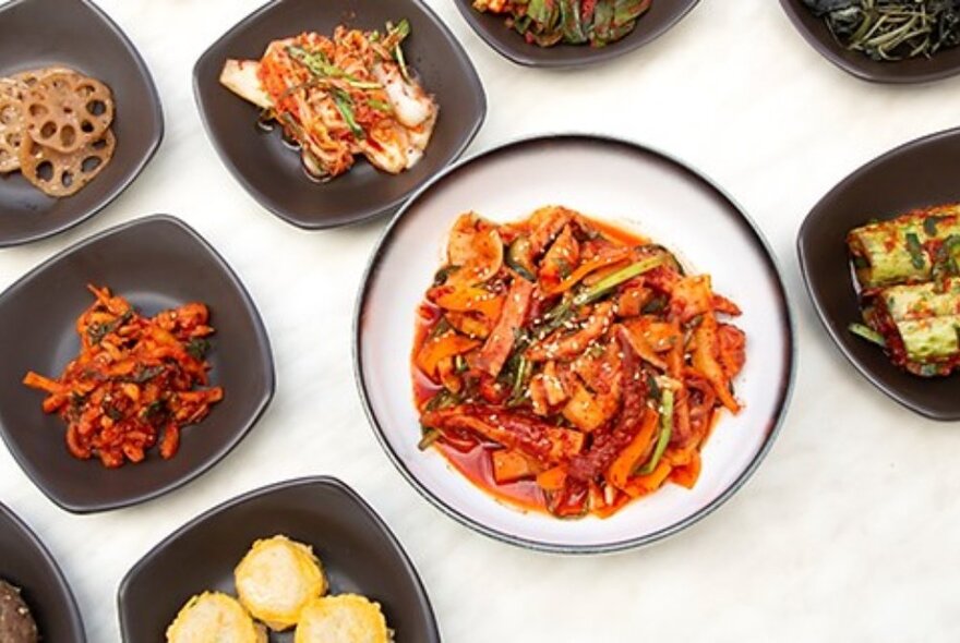 A selection of banchan (small Korean side dishes) surrounding a main plate of a spicy Korean stirfry, on a white surface and seen from above.