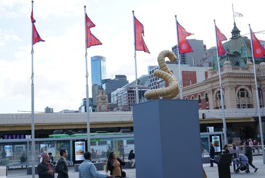 Six Nepalese flags flying on flag poles, with the Flinders Street Station building in the background