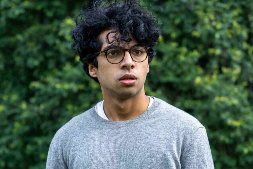 Comedian Rao looking anxious, with dark hair and glasses, against tree backdrop.