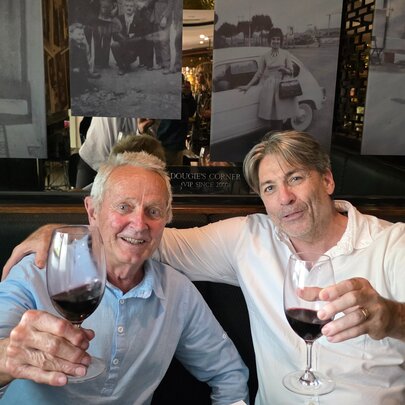 Two men sitting in a restaurant raising a glass or red wine, one with his arm around the other. 