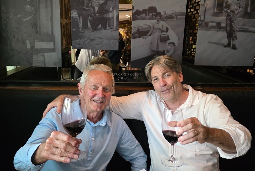 Two men sitting in a restaurant raising a glass or red wine, one with his arm around the other. 