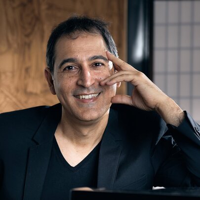Pianist Yitzhak Yedid leaning on a piano with one hand on his face, smiling warmly. 