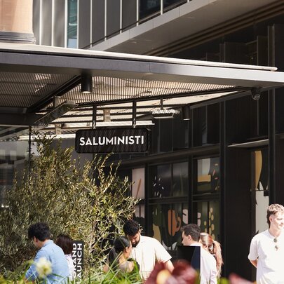 The Saluministi cafe, in Melbourne Quarter, featuring a prominent black sign above an outdoor seating area with people standing outside.