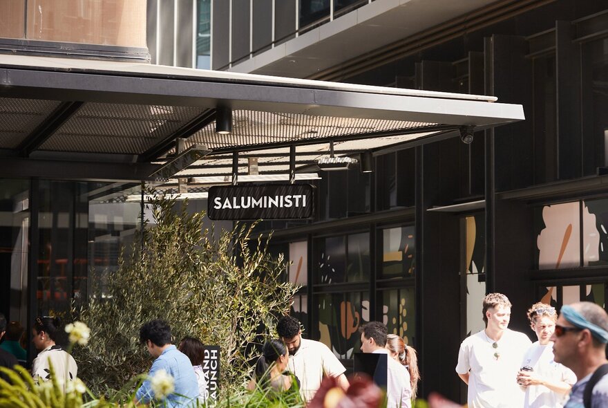 The Saluministi cafe, in Melbourne Quarter, featuring a prominent black sign above an outdoor seating area with people standing outside.