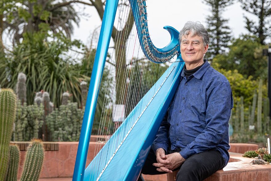 Older man with grey hair wearing a blue shirt, seated outdoors on a stone bench in a garden of succulents, with a large blue harp next to him.