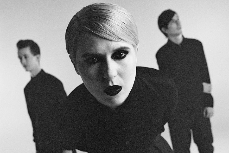 A black and white photograph of the lead singer of the alt rock band Papercity leaning into the camera, with the two other members out of focus behind her.
