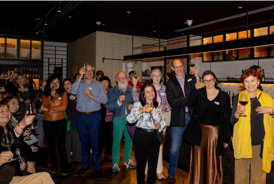 A group of people standing together in a dimly lit indoor venue, smiling and raising their glasses during a social event.
