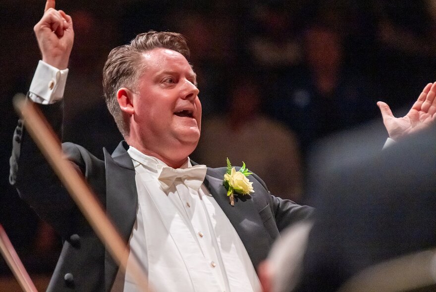 Conductor waving a baton, wearing white tie with buttonhole flower.