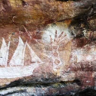 Aboriginal rock art featuring a sailing ship and a hand stencil, on a reddish coloured flat rock surface.