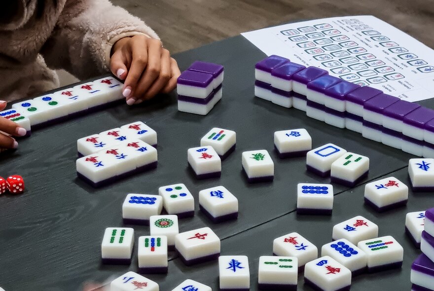 Hands arranging mahjong tiles on a table during a game.