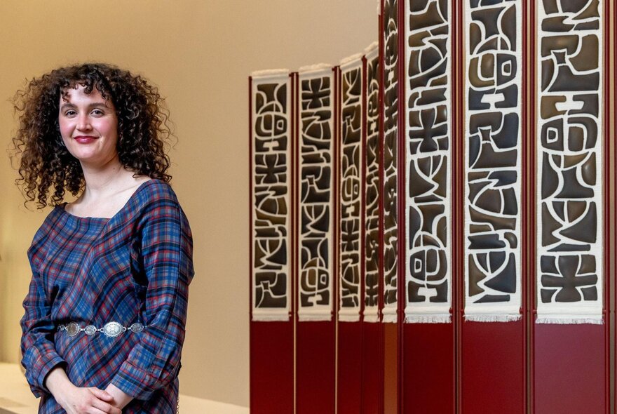A smiling person with curly hair standing in front of a mural like screen display at NGV.