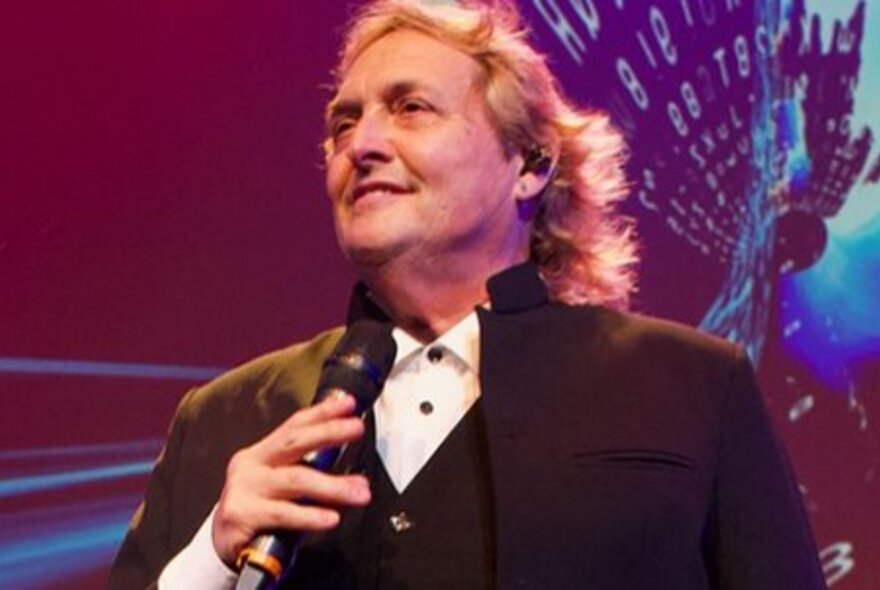 Singer Mike Vee, a middle-aged man wearing a dark suit jacket and white shirt, holding a microphone with wavy blond hair that falls to his shoulders.