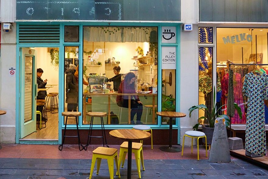 A woman sitting in the window of a blue cafe with seating outside.