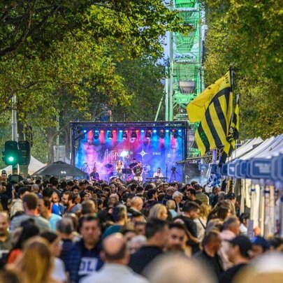 Crowds thronging a street with leafy trees, tent stalls and stage with lighting and live performance as part of the Antipodes Greek Festival.