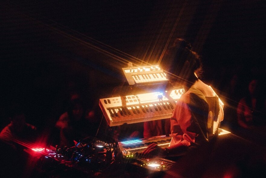 Woman seated at illuminated synthesiser keyboard in the dark.