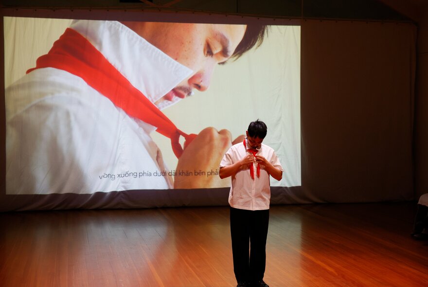 A man facing forwards tying a red tie in a room with a full screen showing a man who is also tying a red tie, but in profile.