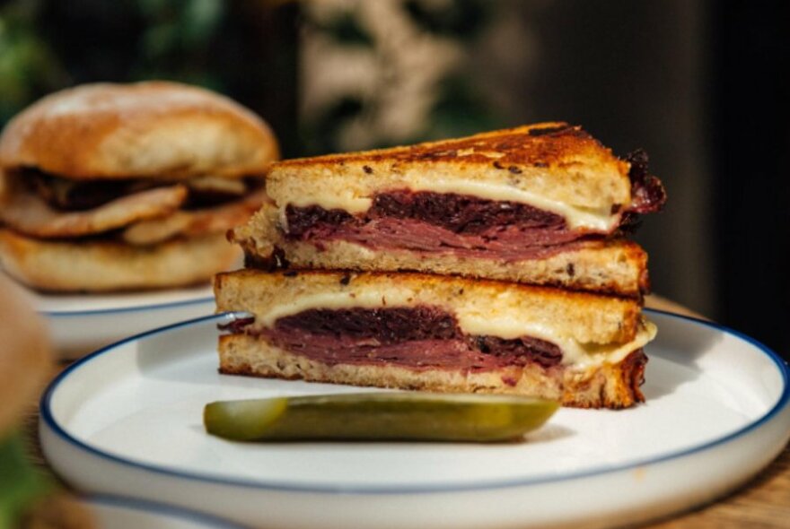 A sliced Reuben toastie on a cafe plate with a pickle. 