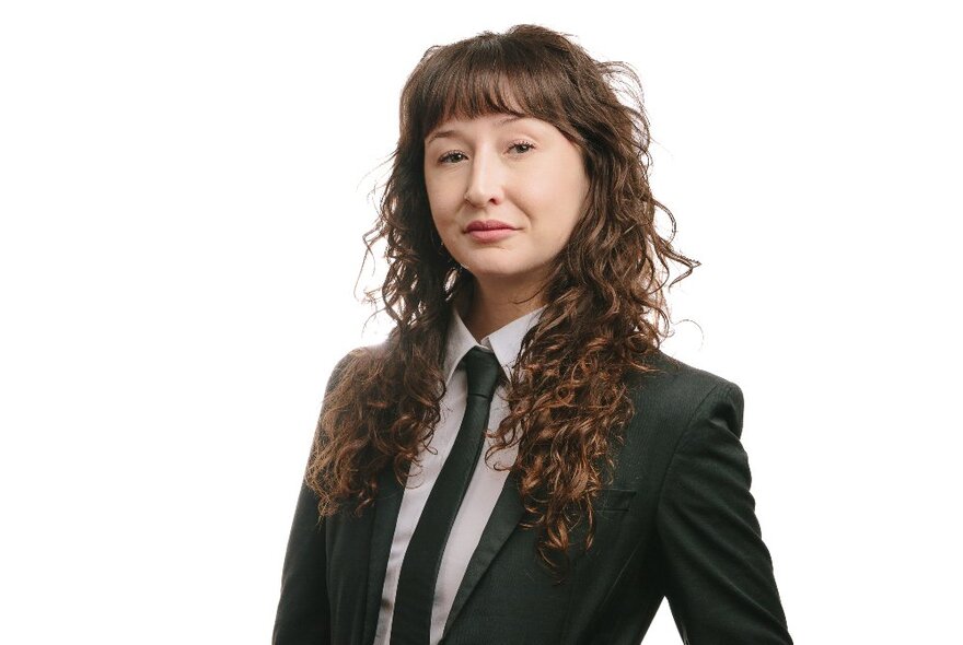 Woman wearing a tie and suit with long curly dark hair, with a sardonic expression.