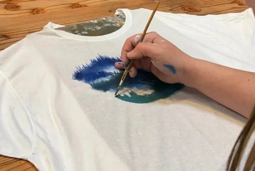 A person is hand-painting a blue design onto a white fabric t-shirt using a fine paintbrush.