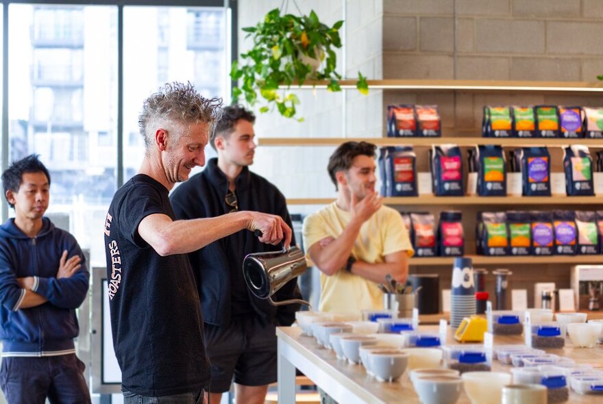 A person hosting a coffee cupping demonstration in a shop, pouring hot water into cups lined up on a counter, with workshop participants watching.