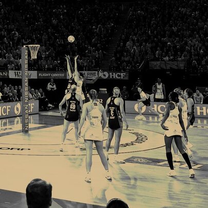 A professional netball match, with players on the court and fans in the stadium seats watching; the image is black and white.