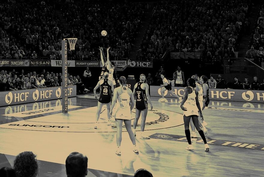 A professional netball match, with players on the court and fans in the stadium seats watching; the image is black and white.