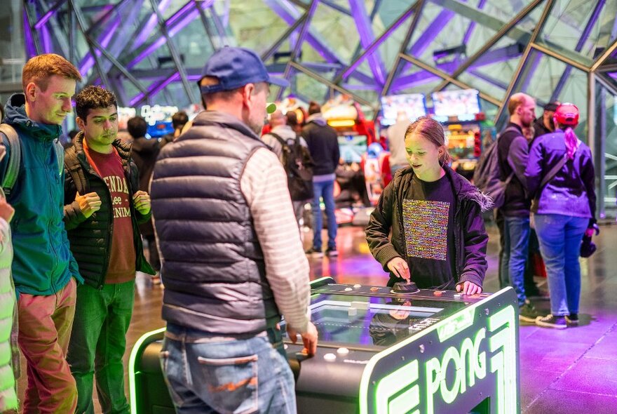 Two people playing an arcade game watched by two others, with other people in the background.