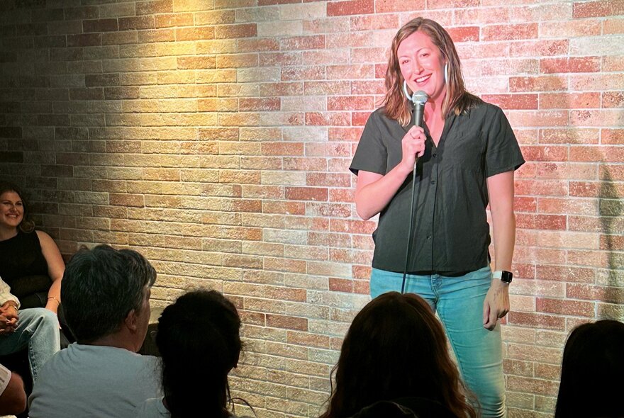 Comedian Celia Pacquola performing stand-up at a small venue with a brick wall background. 